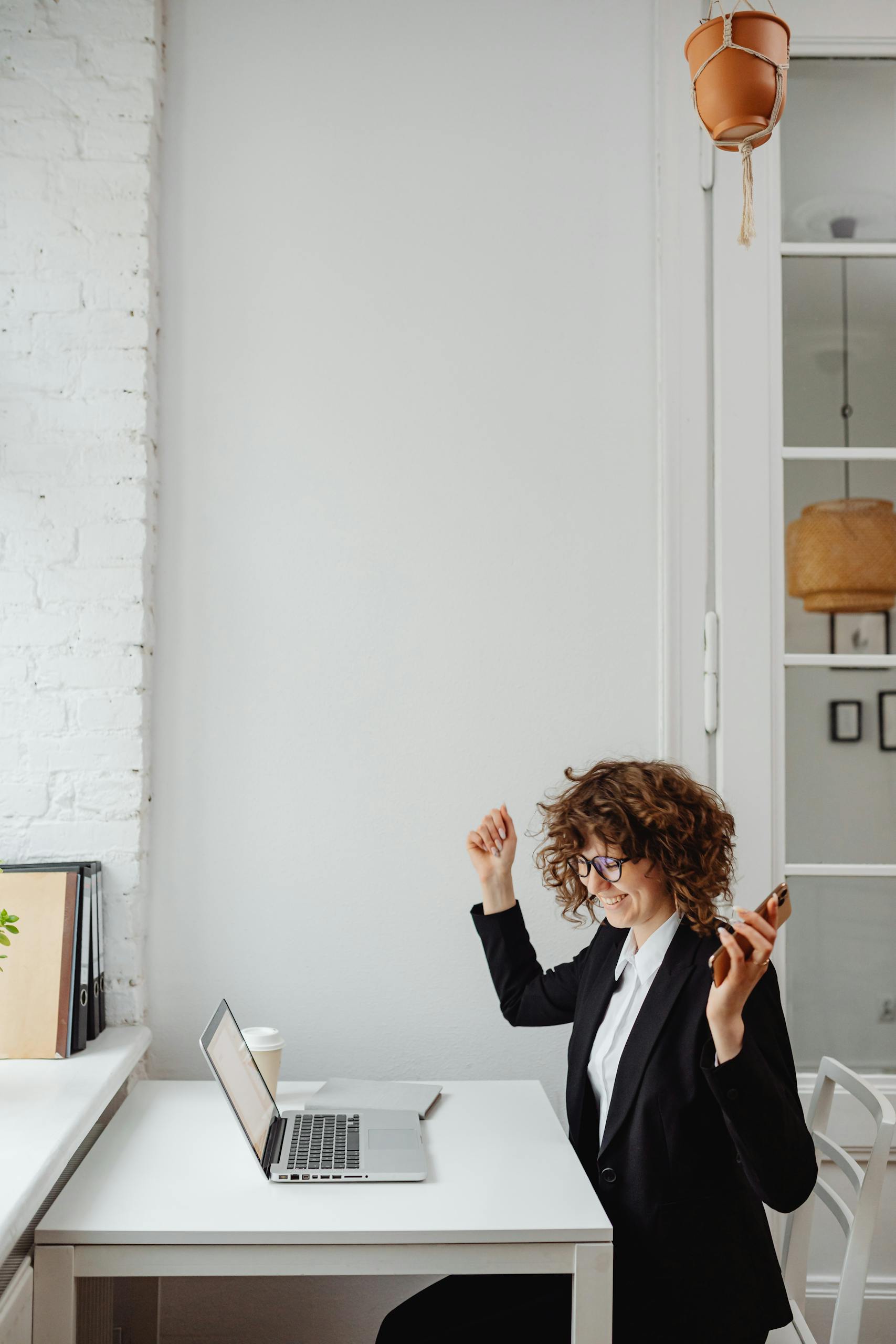 Businesswoman with curly hair celebrates success on phone call in modern office with laptop.